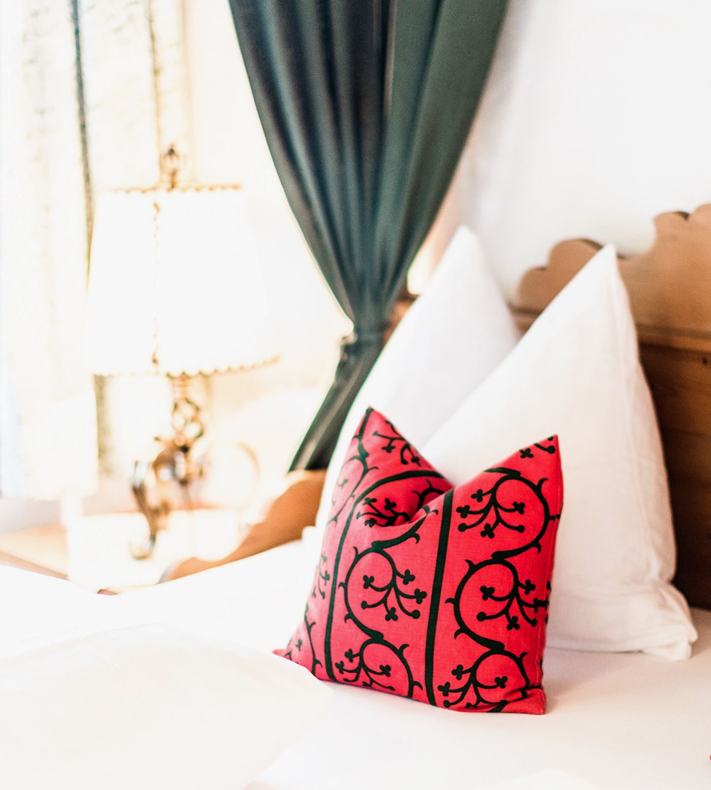 A red decorative pillow with black swirling patterns sits on a white-sheeted bed next to fluffy white pillows.