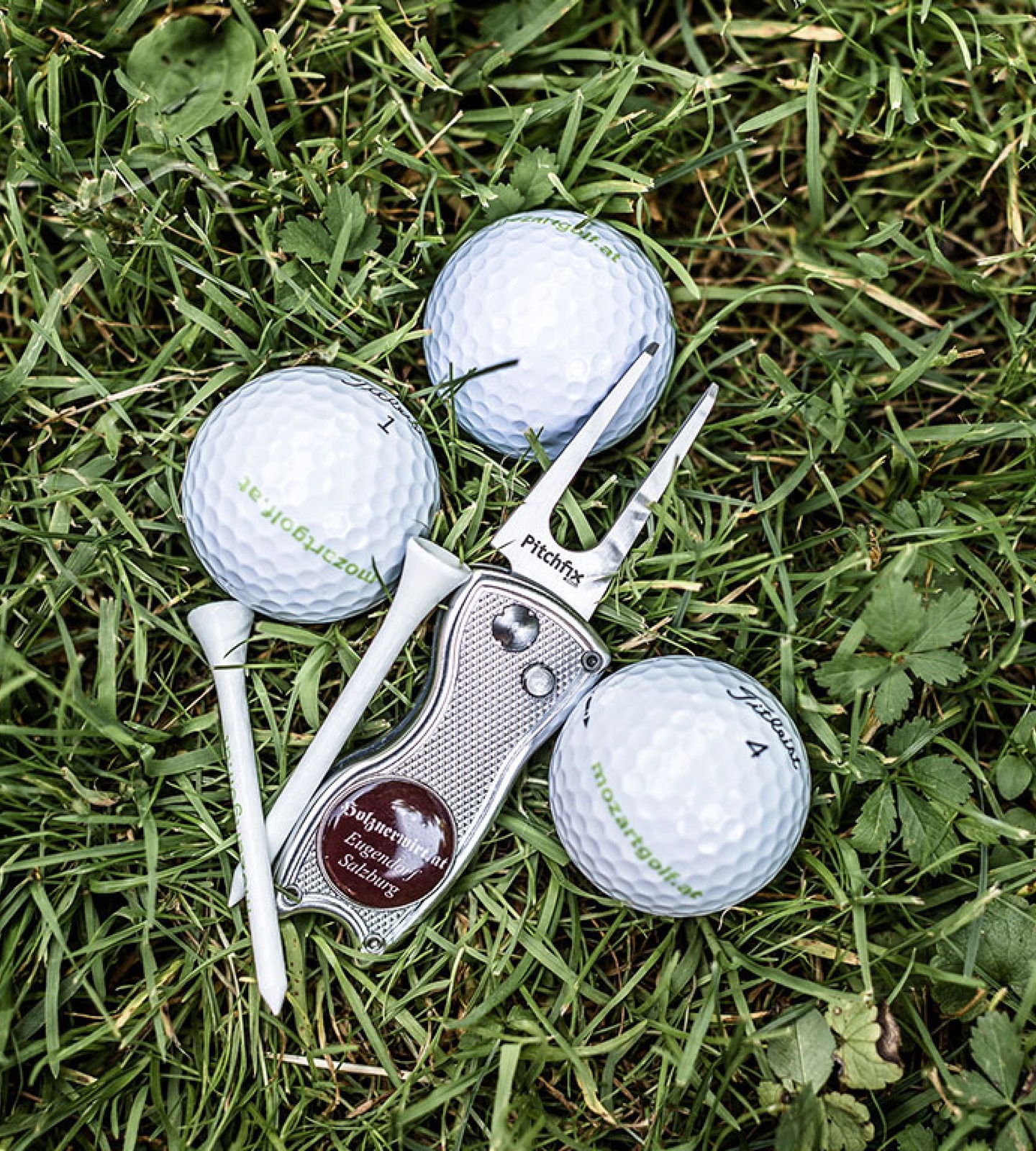 Three Titleist golf balls, two white tees, and a Pitchfix divot tool with a custom logo lie on green grass.
