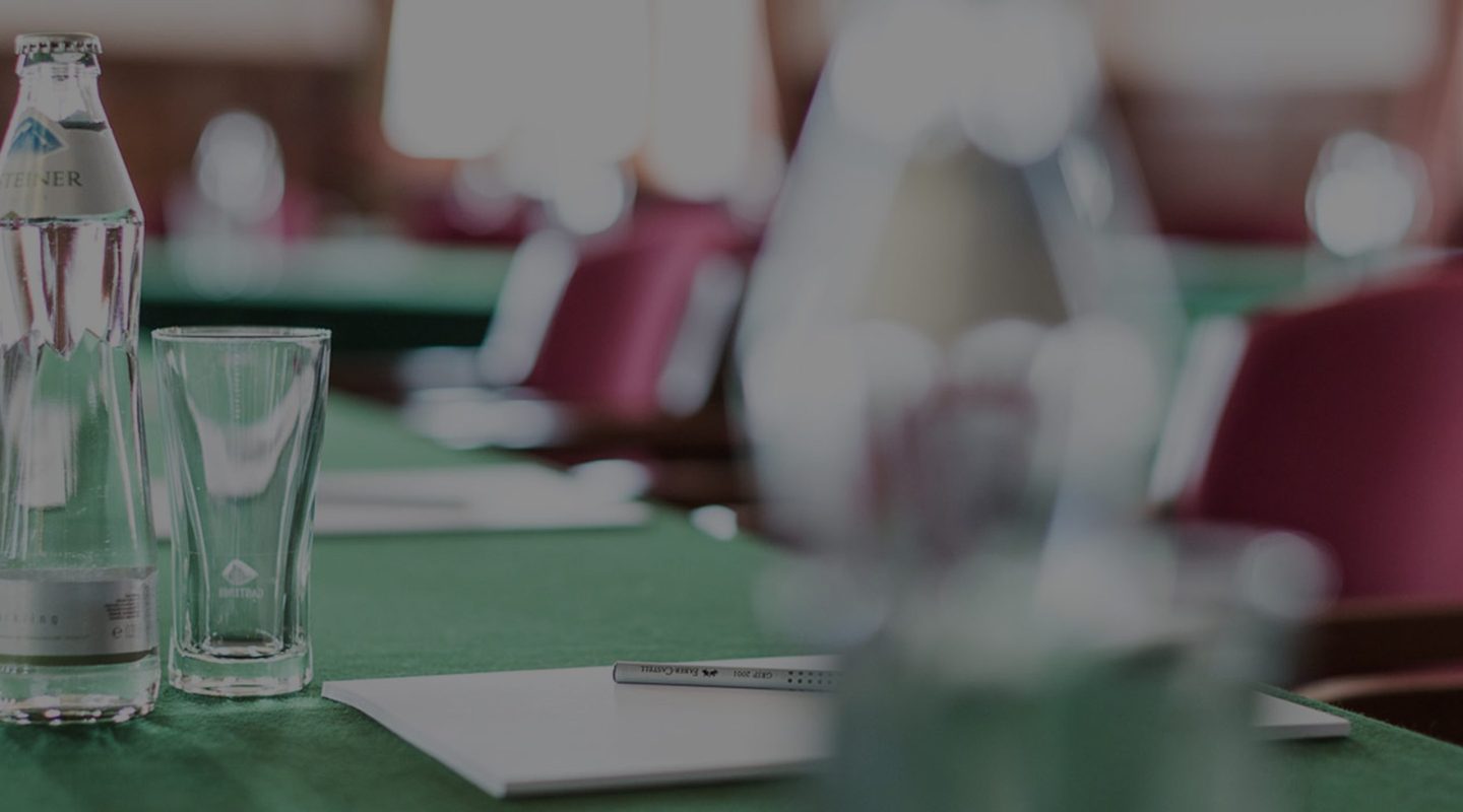 A glass of water, bottle, and notepad on a green table in a conference room with blurred chairs in the background.