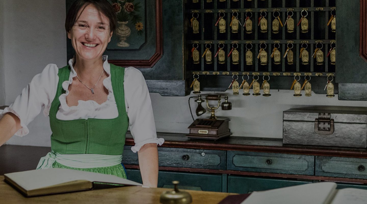 Smiling woman in a green dirndl standing at a rustic hotel reception desk with a key rack and vintage phone behind her.
