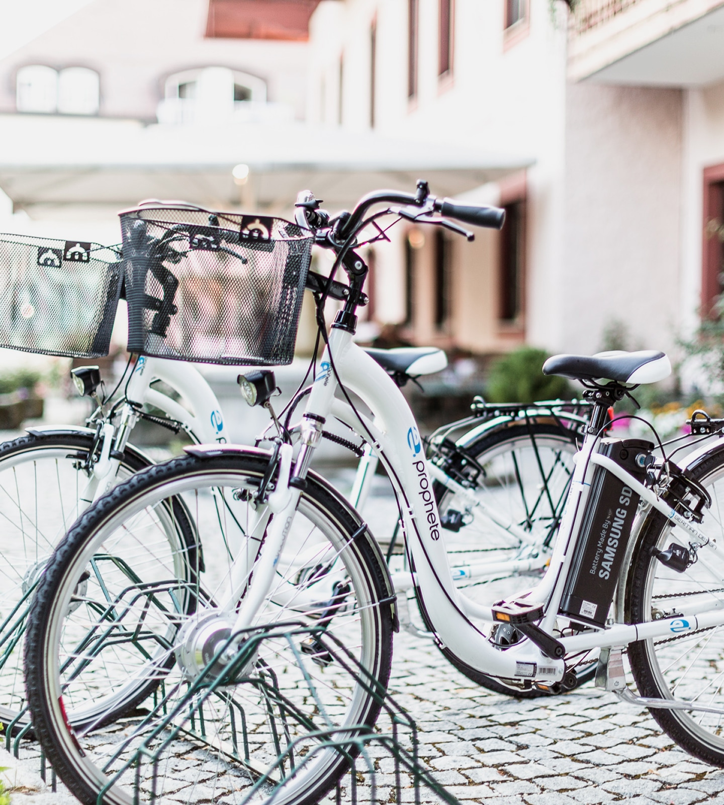 White Prophete electric bicycles with front baskets parked in a rack on a cobblestone street.