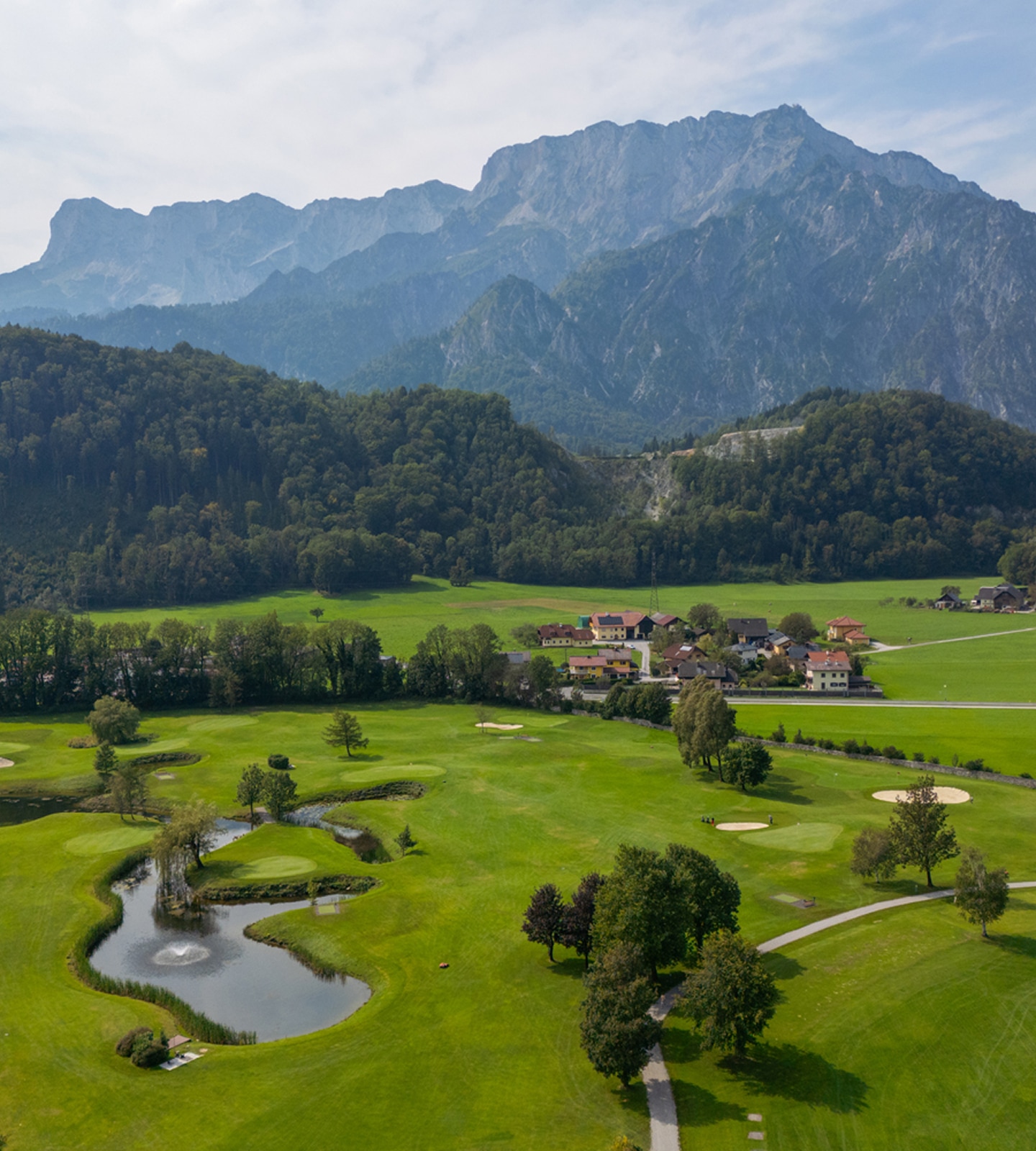 Aerial view of a lush green golf course with water features, a small village, and massive mountains in the background.
