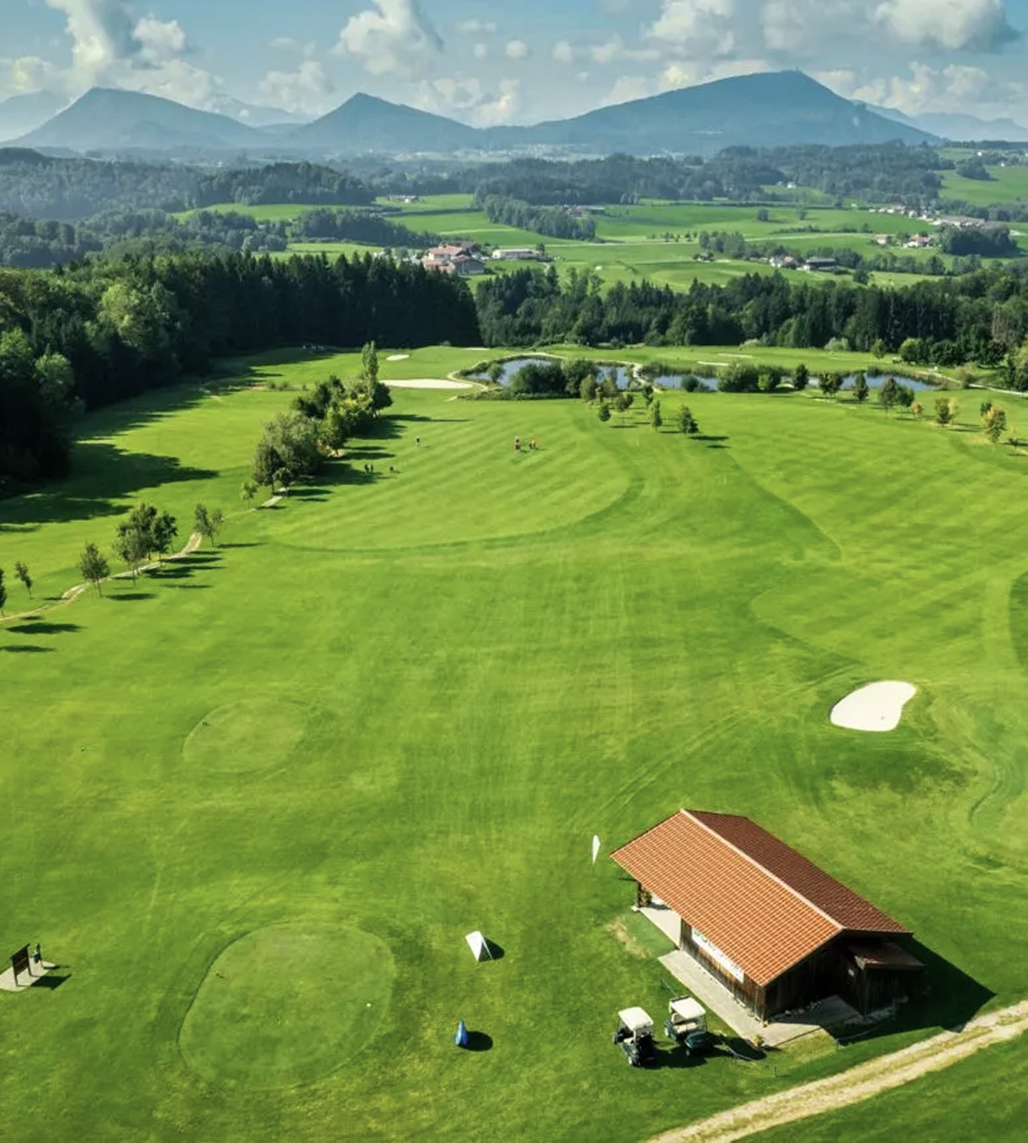 Aerial view of a lush green golf course with a clubhouse, sand bunkers, and mountains in the background.