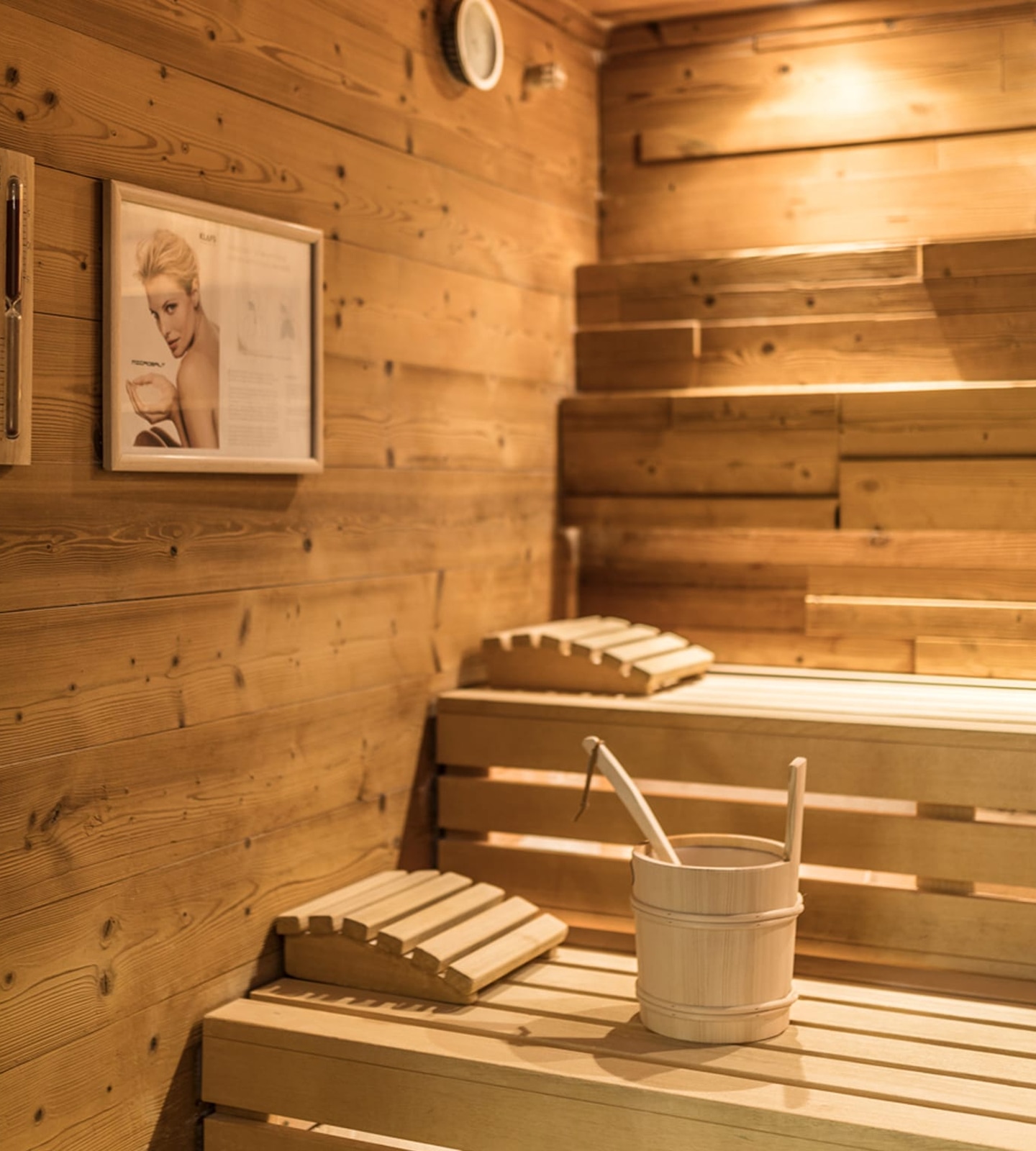 Inside of a wooden sauna room with benches, a bucket with a ladle, headrests, and a framed poster on the wall.