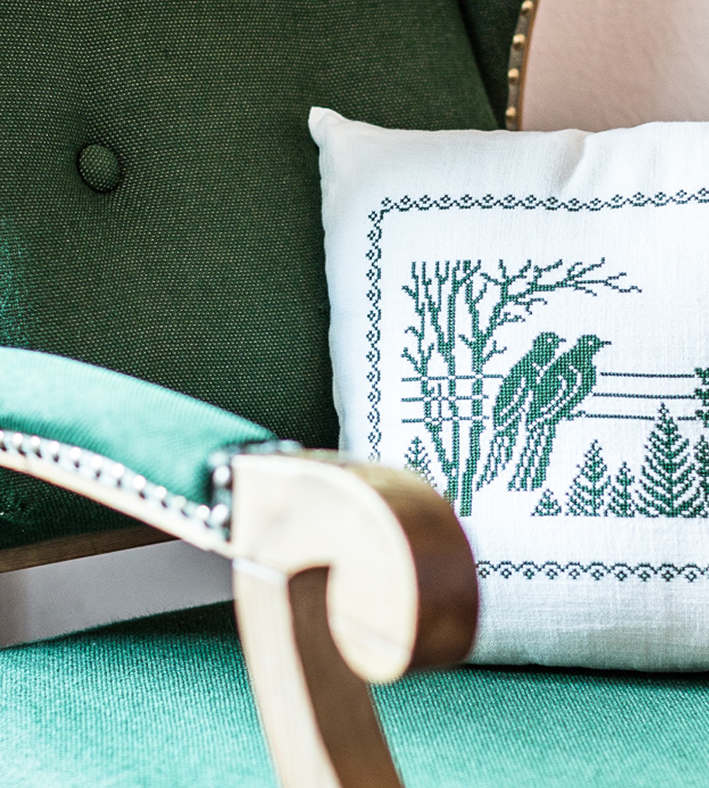 White cushion with green cross-stitch design of birds and trees on a green upholstered armchair.