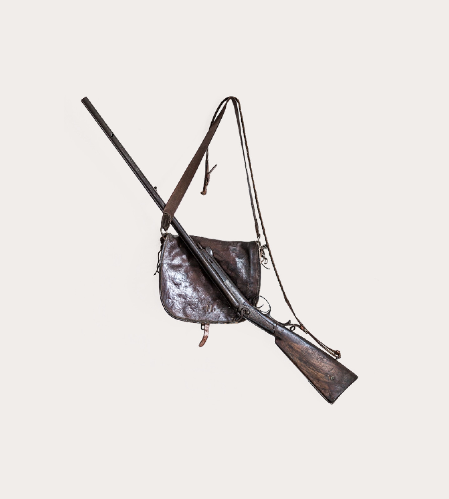 An antique long gun rifle laid across a weathered brown leather shoulder bag on a plain background.