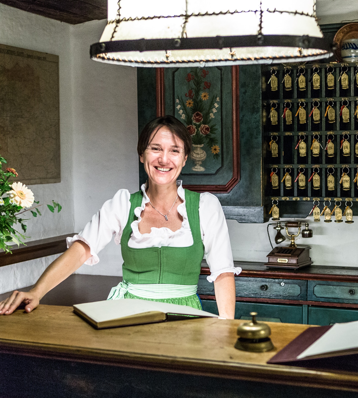 Smiling woman in a green dirndl standing at a rustic hotel reception desk with a key rack behind her.