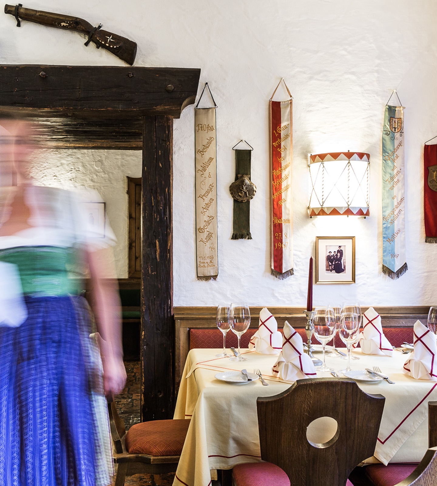 A blurred waitress in a dirndl walks past a set dining table with banners on the white wall in a traditional restaurant.