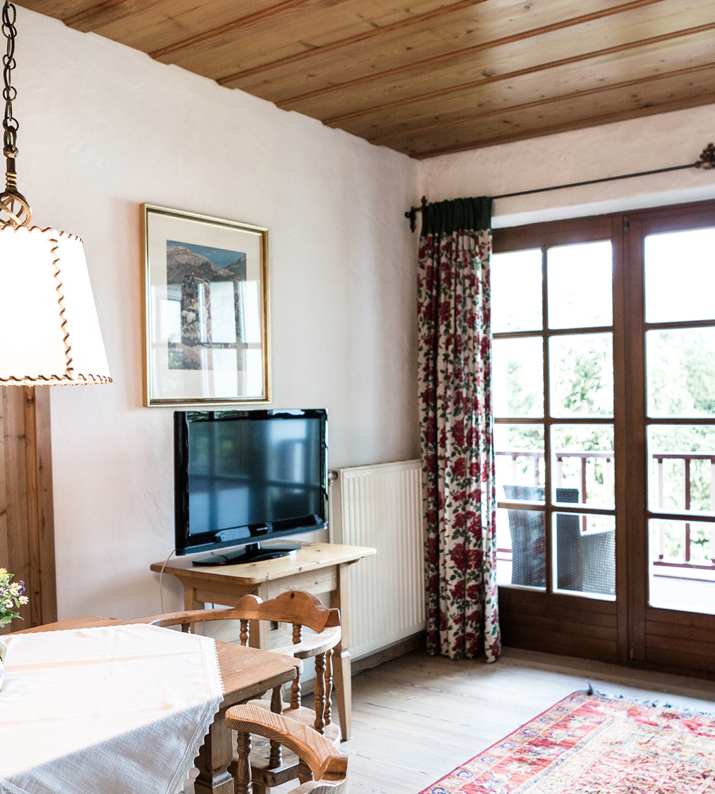 Cozy rustic living area with wooden ceiling, a flat-screen TV on a table, floral curtains, and a balcony door.
