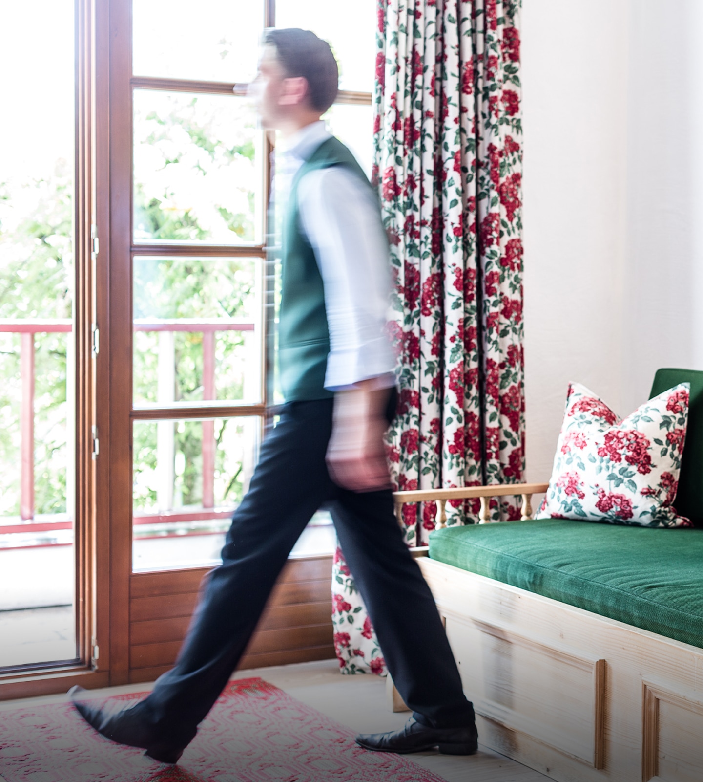 A blurred motion shot of a man in a green vest walking past a window in a room with a green bench.