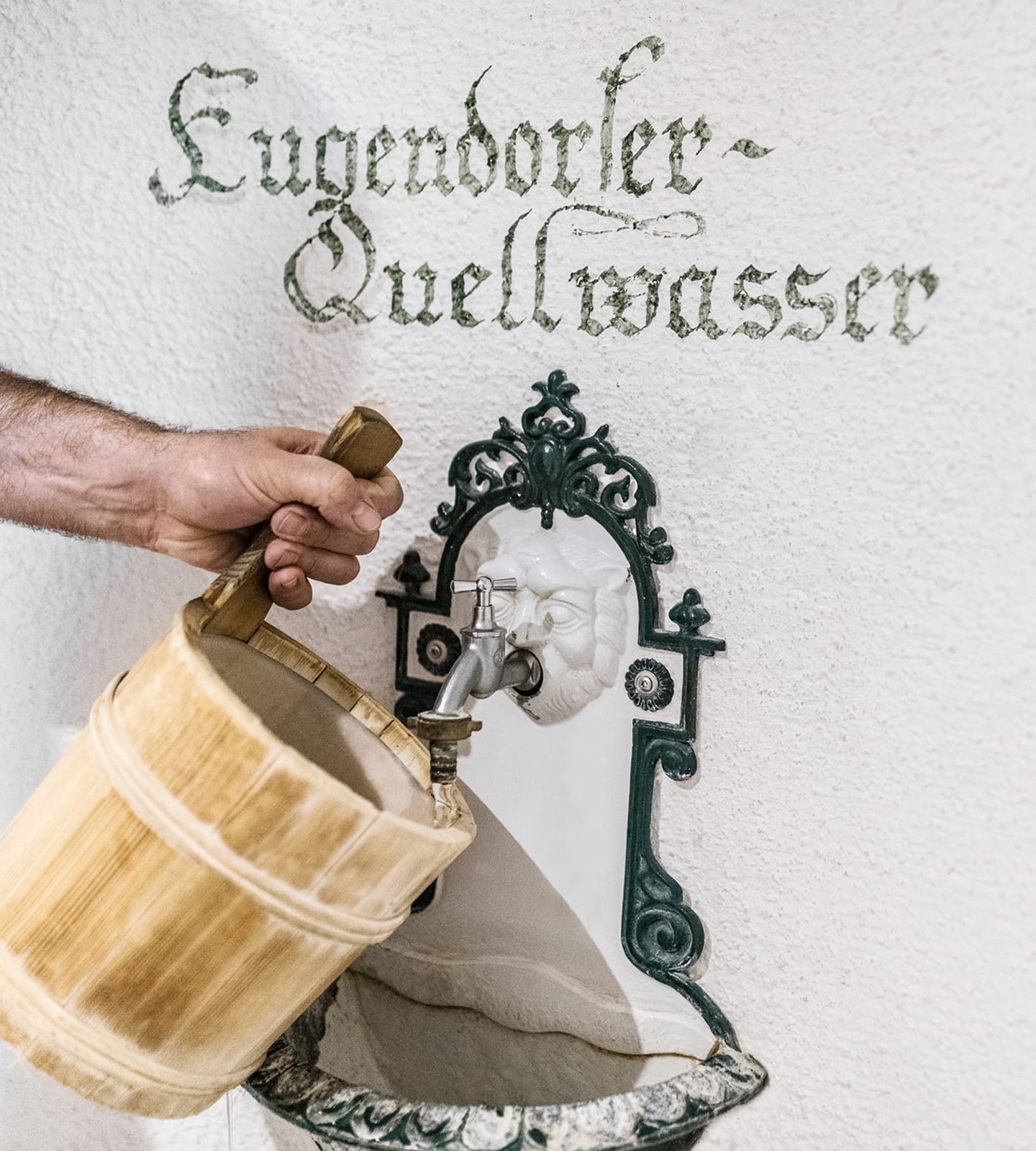 A hand holds a wooden bucket under a decorative wall fountain with text reading "Eugendorfer Quellwasser."