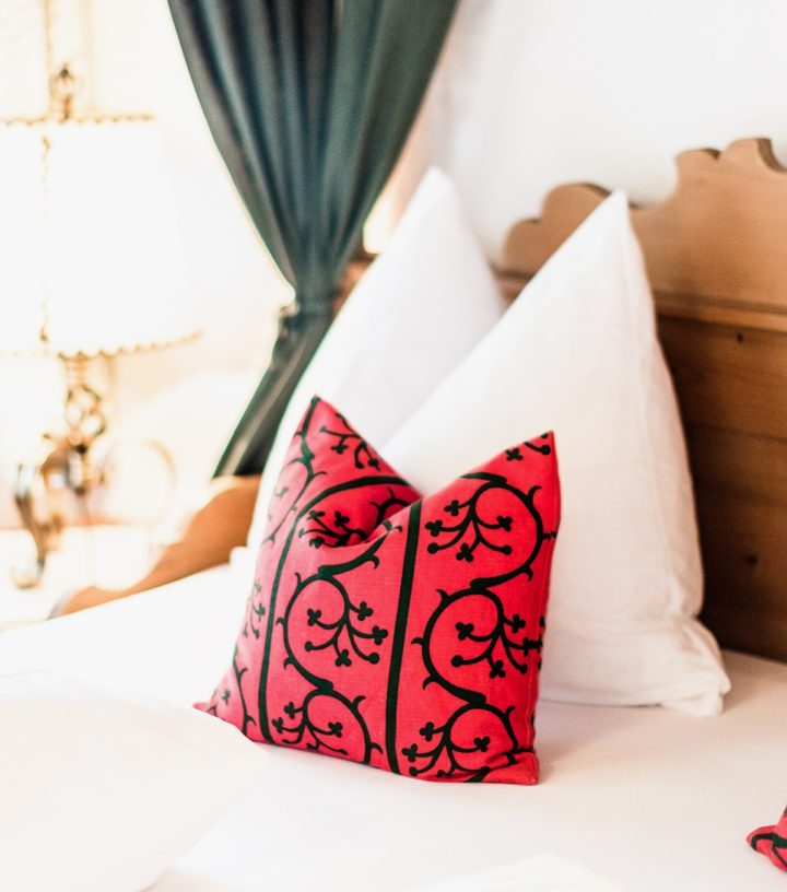 Cozy bed with wooden headboard, large white pillows, and red pillows with black scroll patterns next to a window.