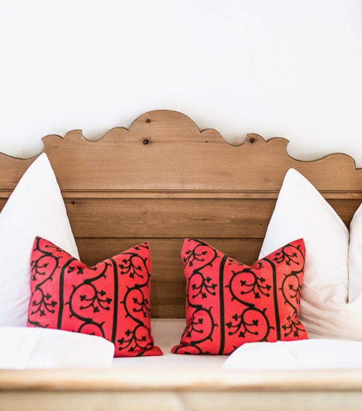 Wooden bed with a carved headboard, white pillows, red accent pillows, and dark green curtains on either side.