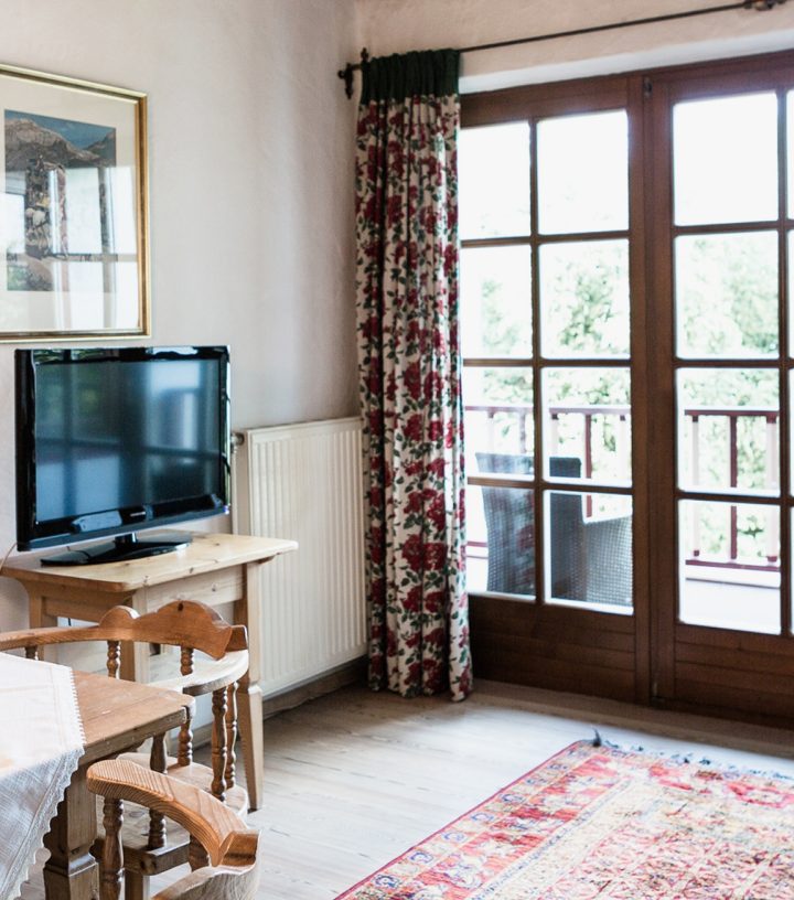 Cozy living area with a wooden table, TV on a stand, patterned rug, and large french doors leading to a balcony.