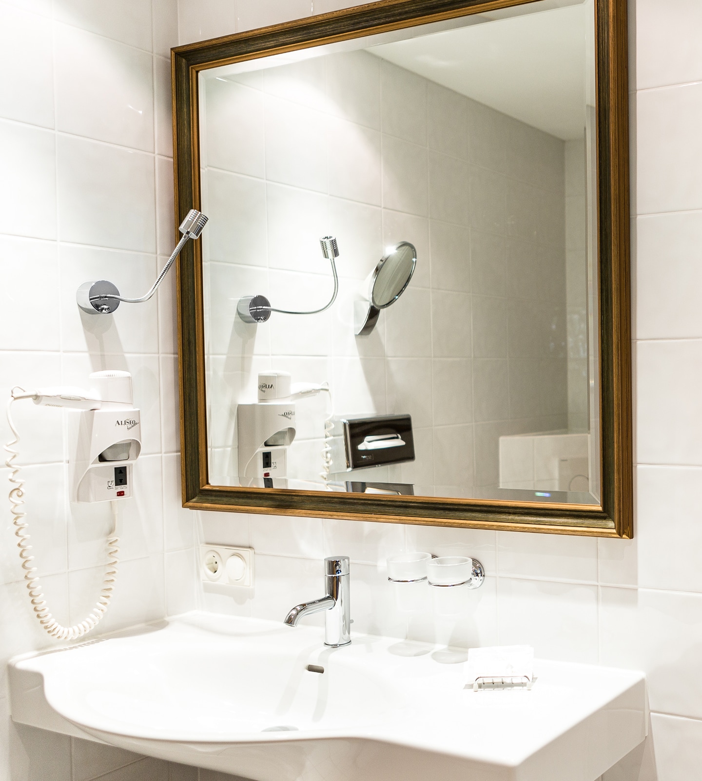 Modern bathroom sink area with a white basin, chrome tap, wall-mounted hair dryer, and a large gold-framed mirror.