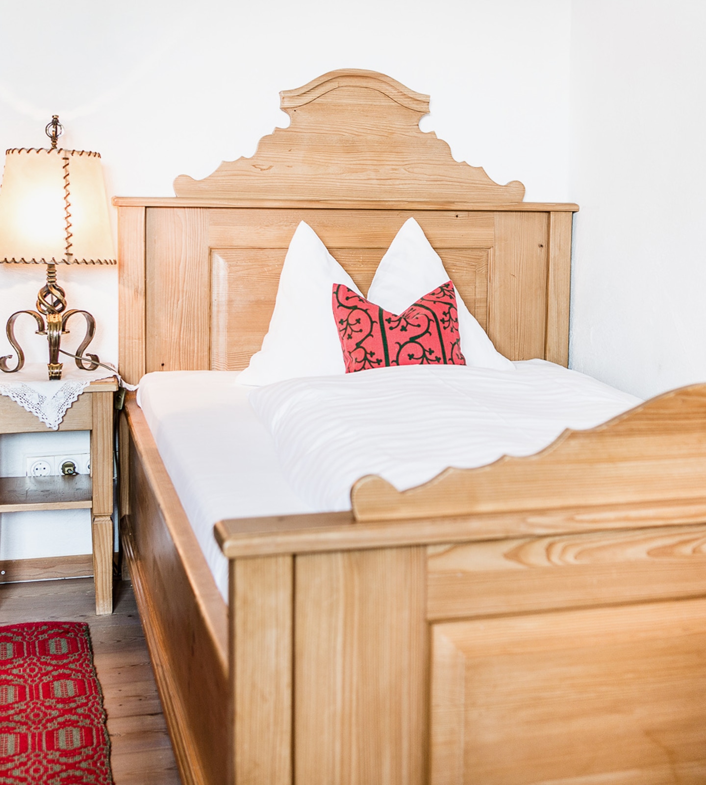 Rustic wooden bed with white linens, two pointed pillows, and a small red accent pillow next to a bedside lamp.