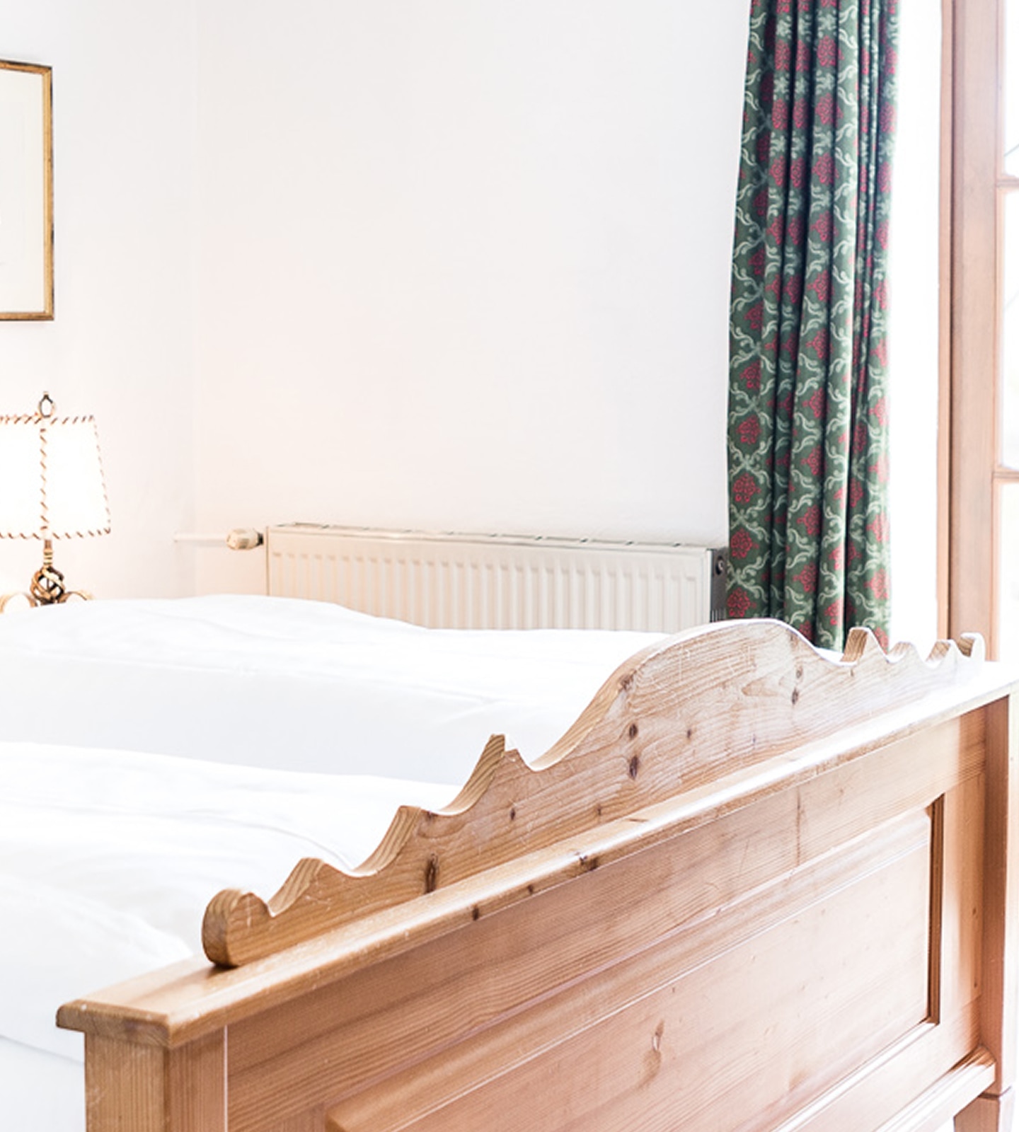 A wooden footboard of a bed with white sheets in a bedroom featuring patterned green curtains.