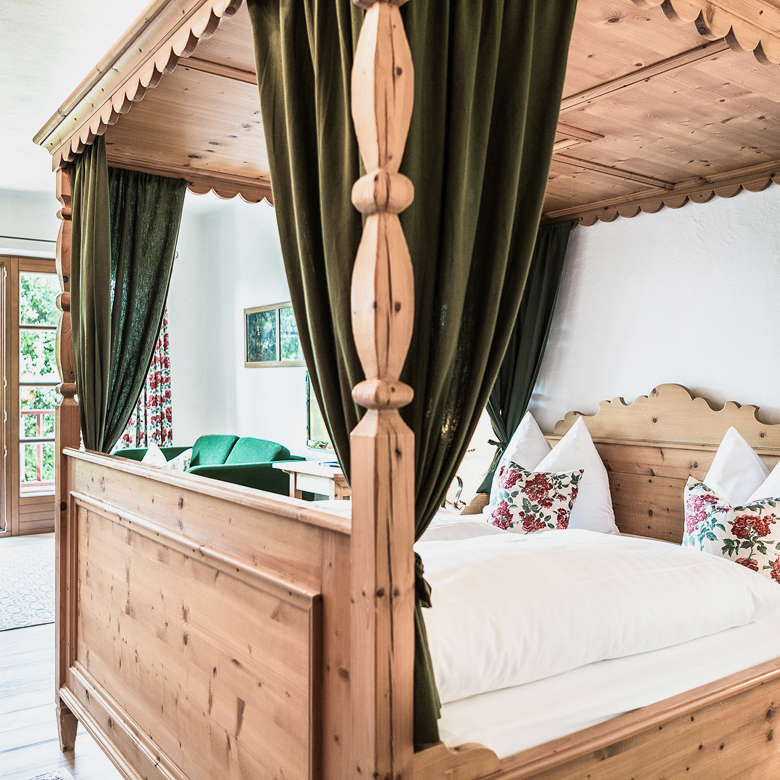 Rustic wooden four-poster bed with olive green curtains and floral pillows in a cozy alpine bedroom.
