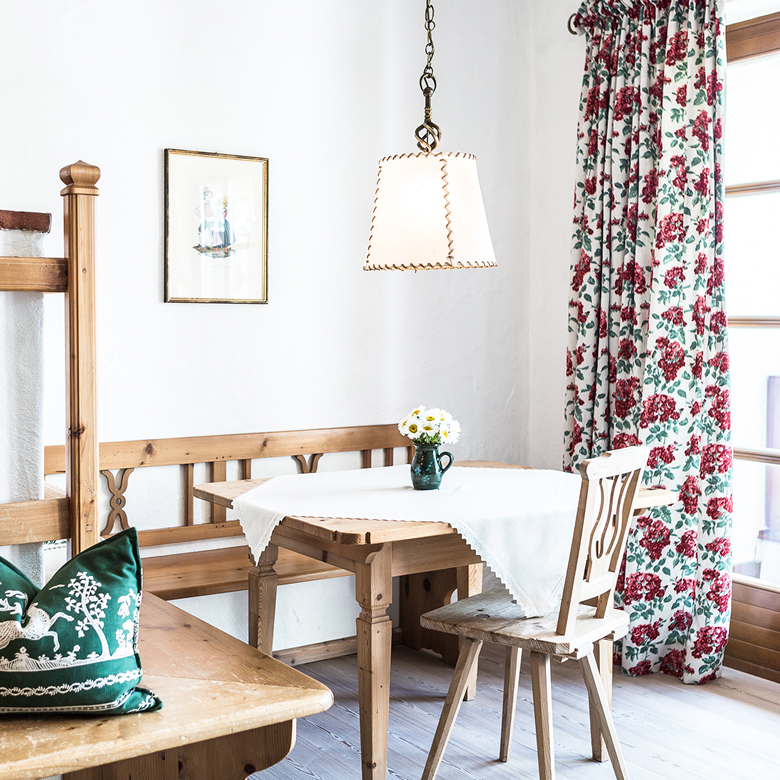Rustic dining area with a wooden table, bench, chair, floral curtains, and a hanging lamp.
