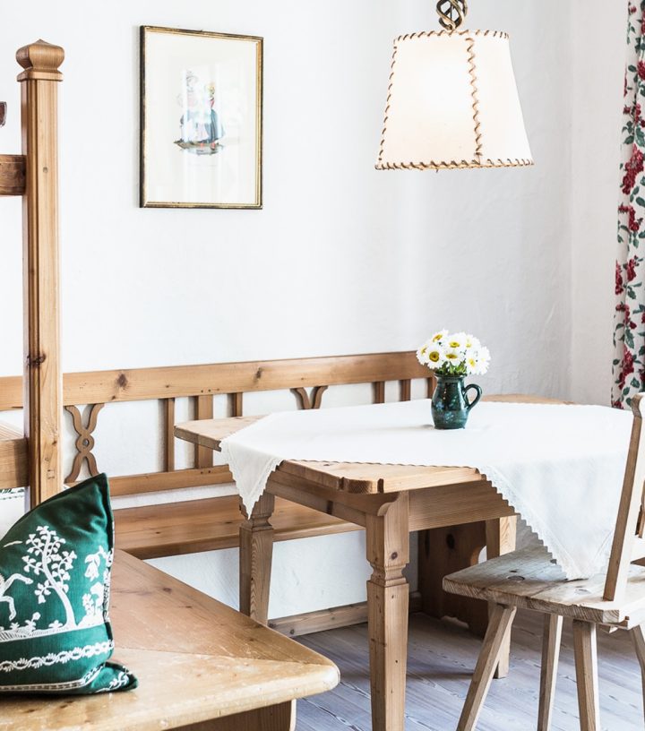 Rustic wooden dining nook with a white tablecloth, hanging lamp, floral curtains, and a green patterned cushion.