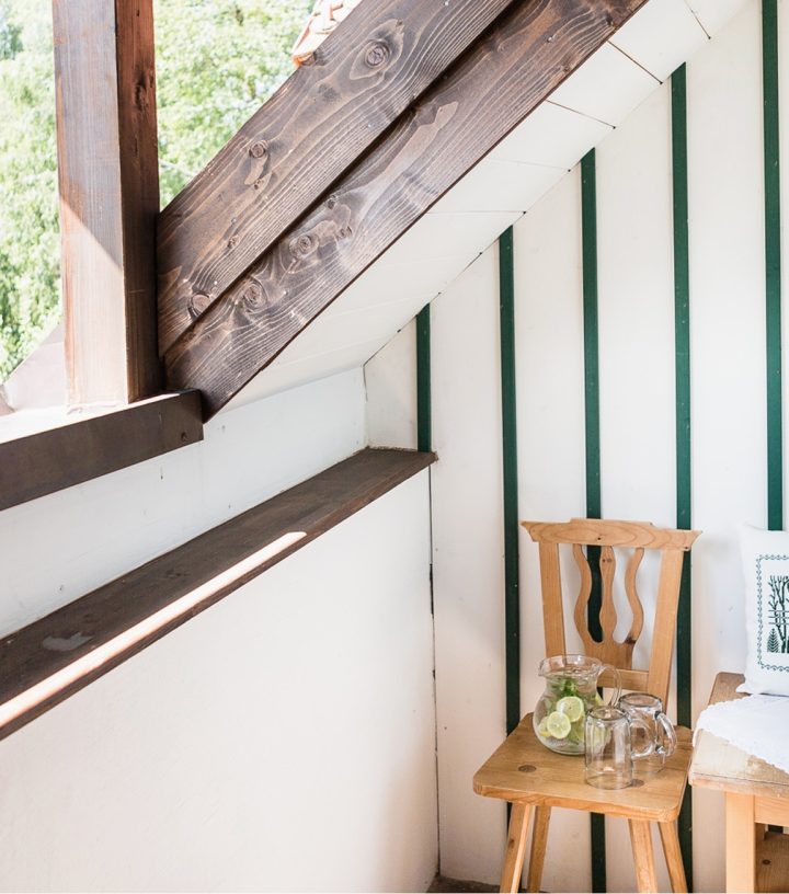 Cozy wooden balcony corner with a small table, chair, flowers, and a pitcher of water overlooking green trees.