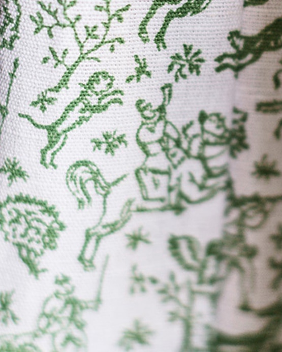 Close-up of white woven fabric with green embroidered patterns of figures, horses, and trees.