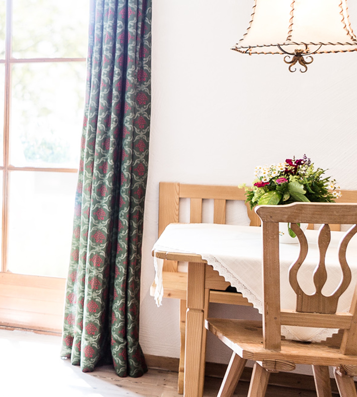 Rustic wooden dining table and chairs with patterned curtains and a floral bouquet centerpiece.