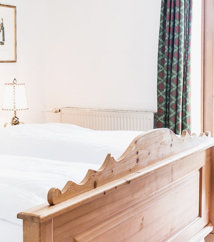 Rustic wooden double bed with white bedding, pink patterned pillows, and a large window in a bright room.