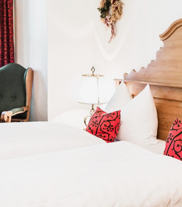 A rustic wooden bed with white linens and red patterned pillows sits in a cozy room next to a green armchair.