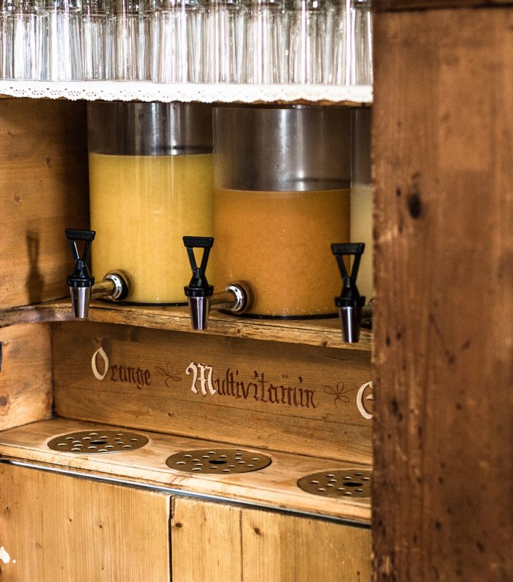 Wooden drink station with dispensers labeled "Orange" and "Multivitamin" containing yellow and orange juices.