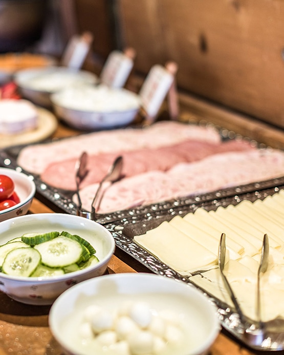 A breakfast buffet with platters of sliced cheese and ham, alongside bowls of sliced cucumbers and cheese balls.