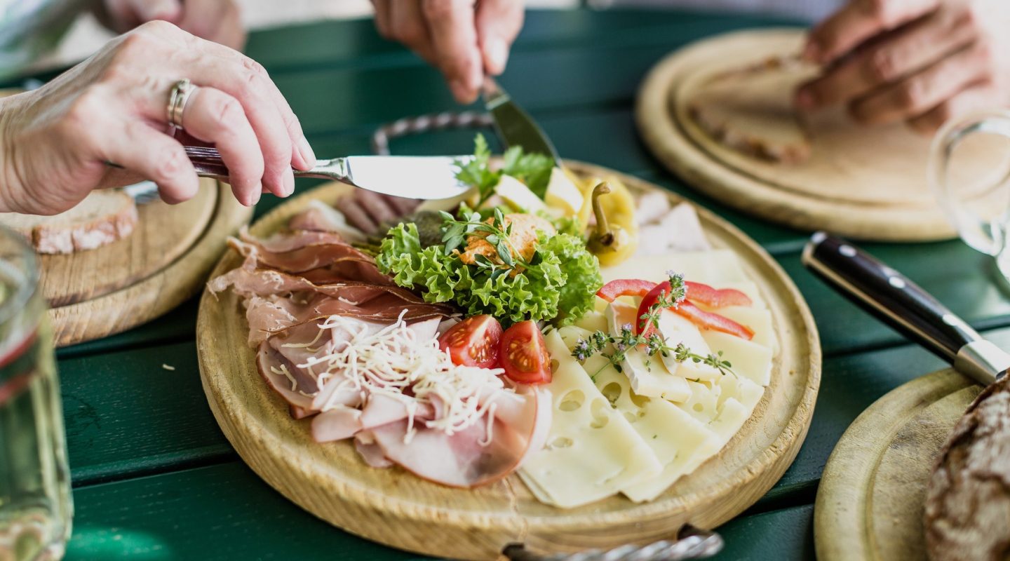 Close-up of hands using a knife to serve a wooden platter filled with various meats, cheeses, and lettuce.