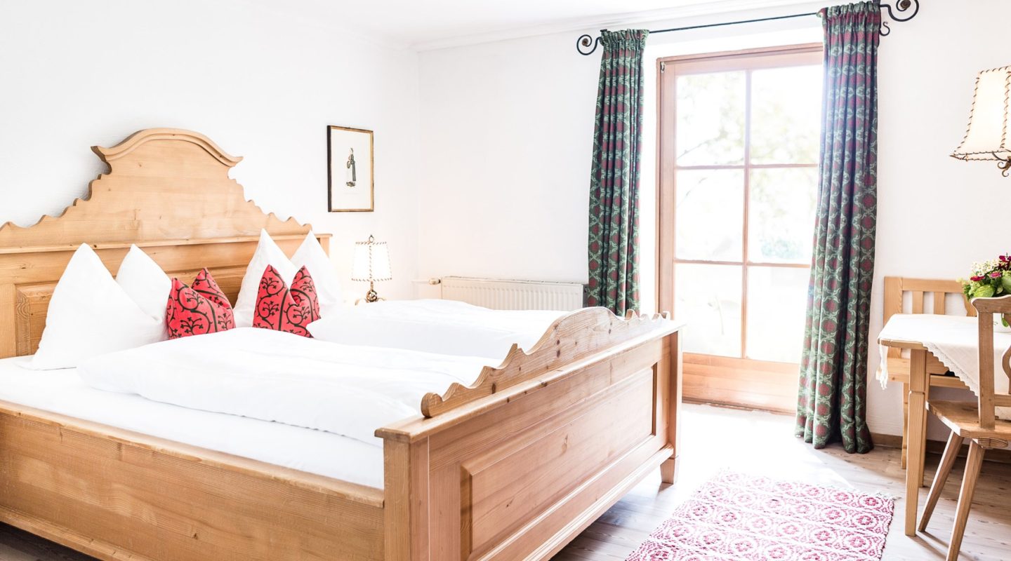 Rustic wooden double bed with white bedding, red patterned pillows, and a wooden table set by a glass door.