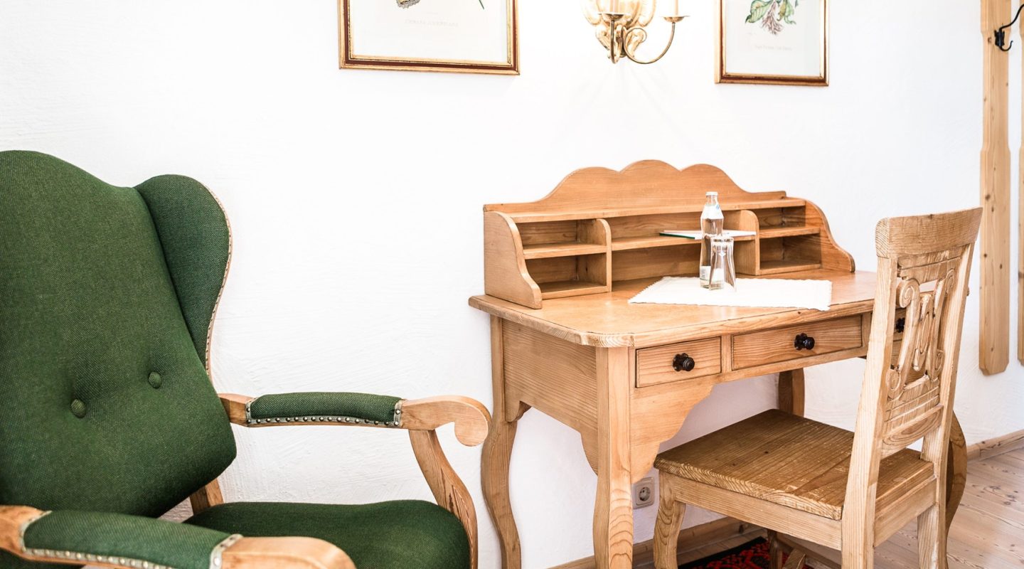 A rustic wooden writing desk and chair next to a green upholstered armchair against a white wall.