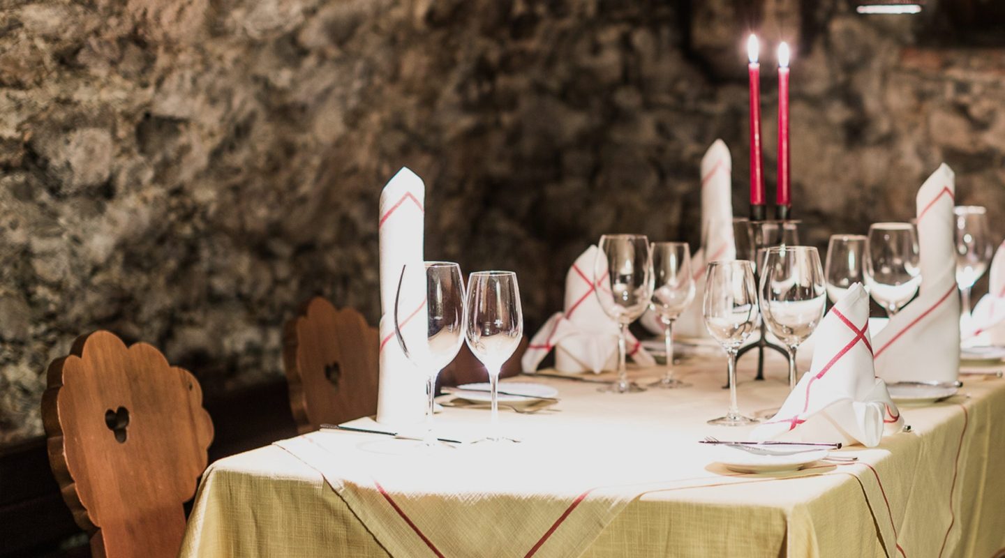 Set dining table in a rustic stone wall room with wine glasses, white napkins, and lit red candles.