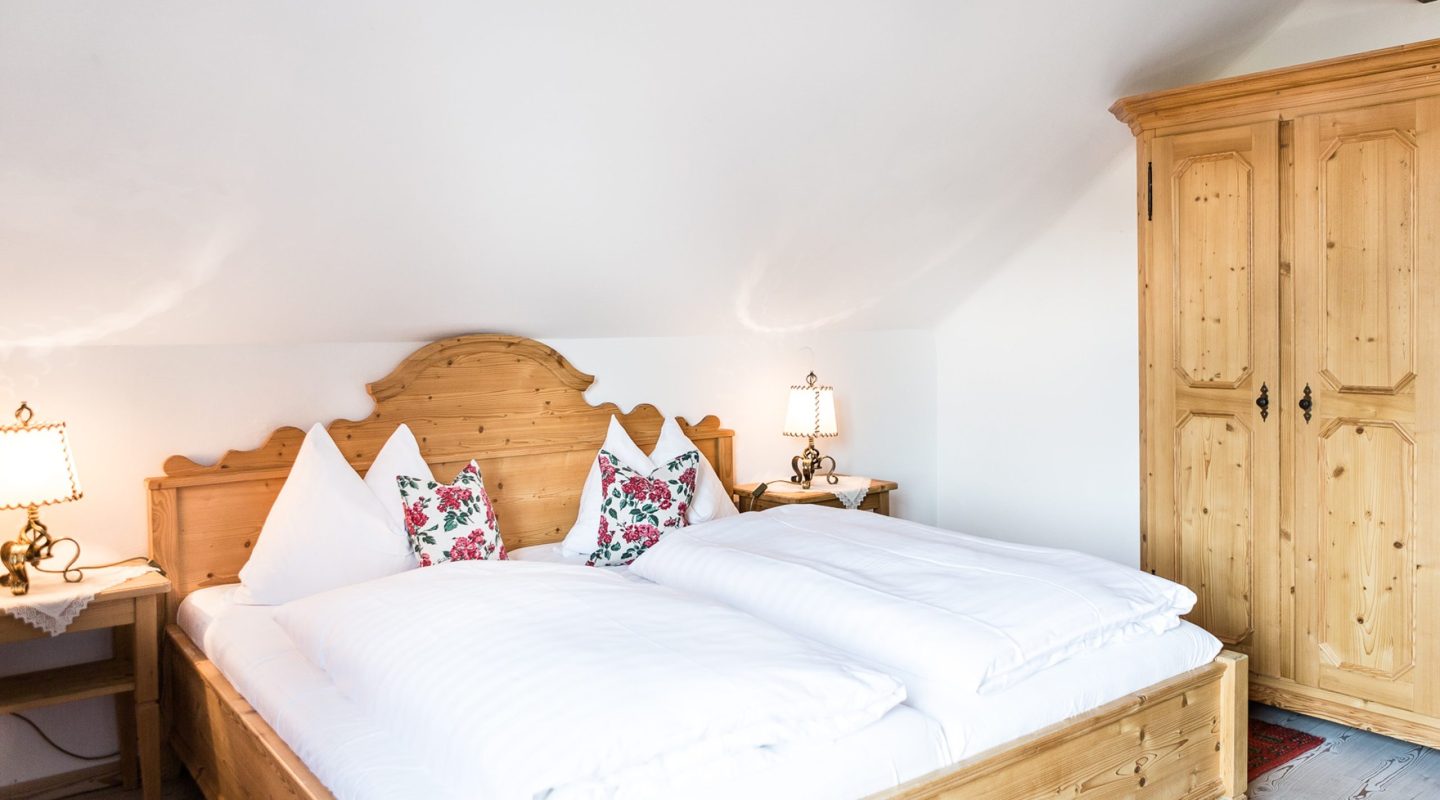 Cozy attic bedroom with a wooden double bed, white duvets, floral pillows, matching side tables, and a wooden wardrobe.