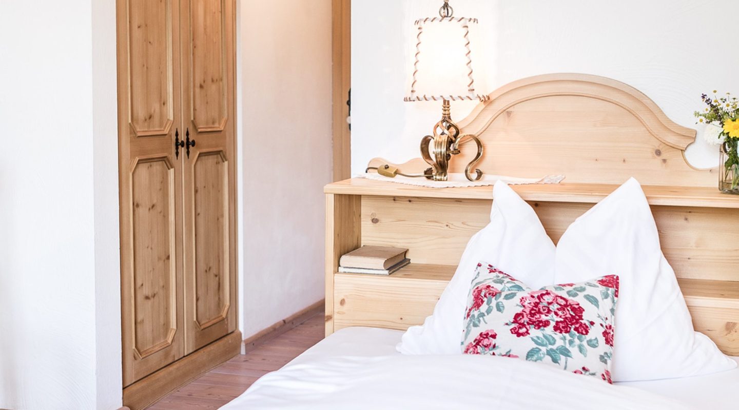 Cozy bedroom with light wood furniture, white bedding, a floral pillow, and an ornate table lamp on the headboard shelf.