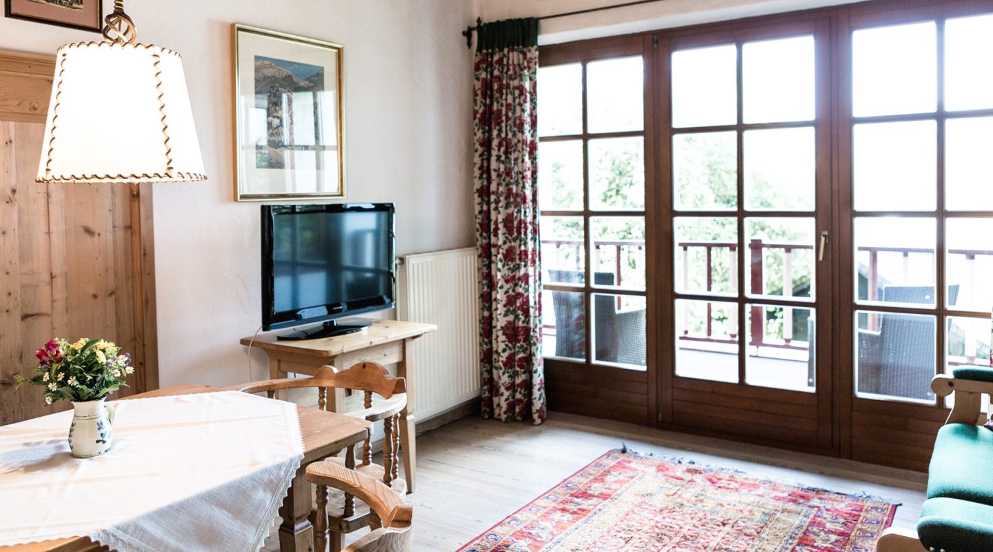 Cozy rustic living room with a dining table, television, patterned rug, and large french doors opening to a balcony.