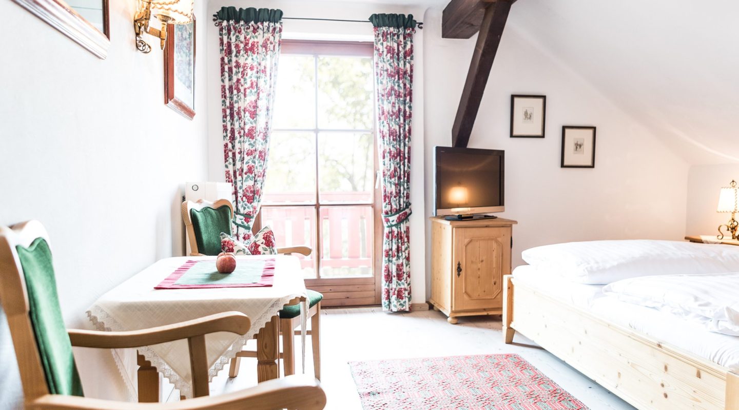 Cozy attic bedroom with a wooden bed, dining table, floral curtains, and a TV on a wooden cabinet.