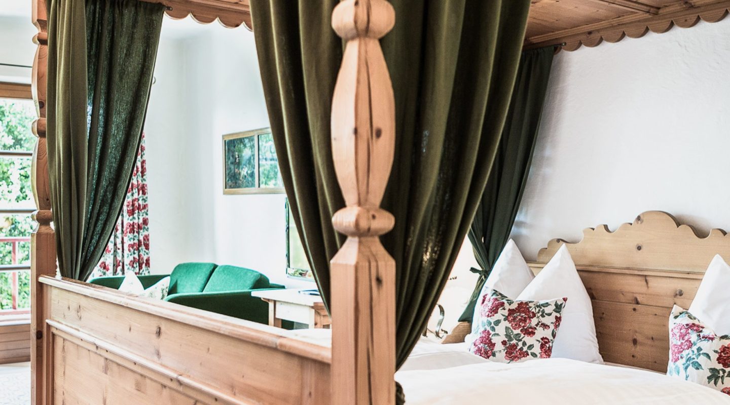 Rustic wooden four-poster bed with green drapes, floral pillows, and a green sofa visible in the background.