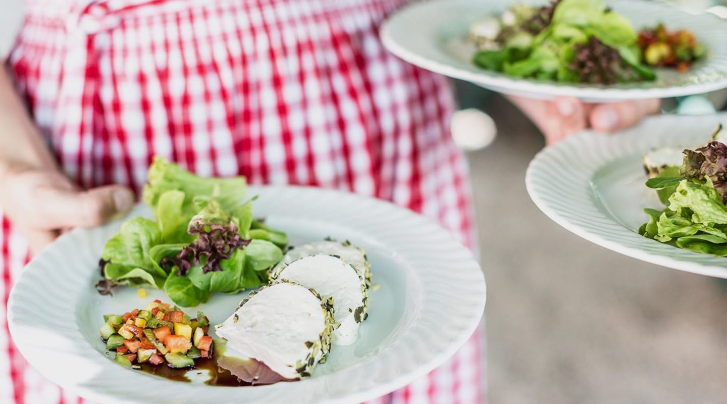 A server holding plates of food featuring sliced goat cheese, fresh green salad, and a finely diced vegetable relish.