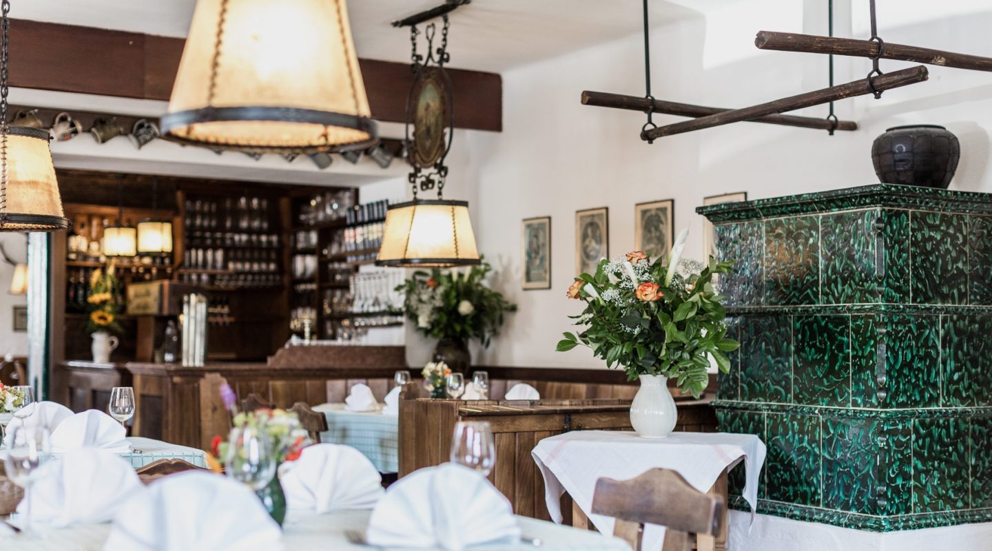 Rustic restaurant interior with set tables, warm hanging lamps, and a decorative green tiled stove.