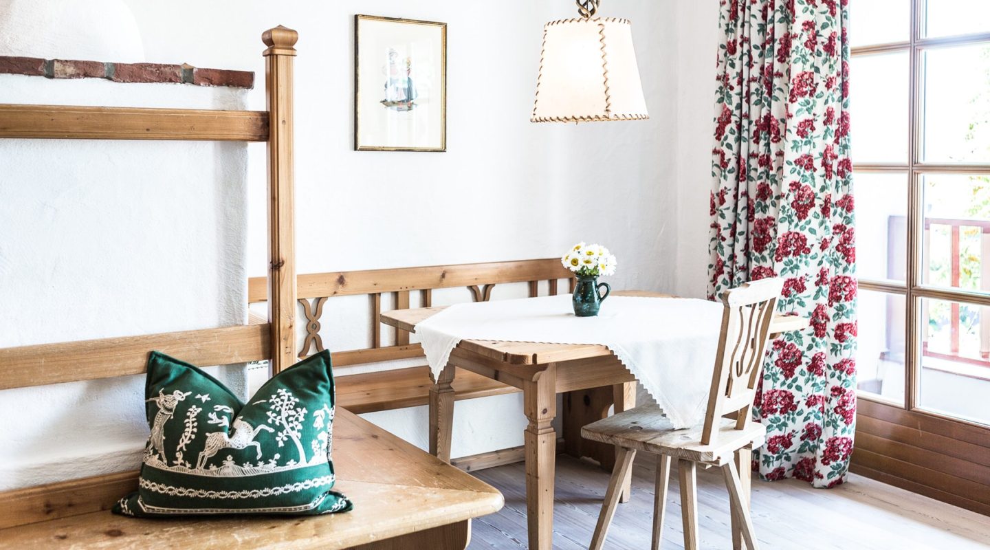 Rustic dining area with a wooden table, bench, chair, floral curtains, and a green decorative pillow.