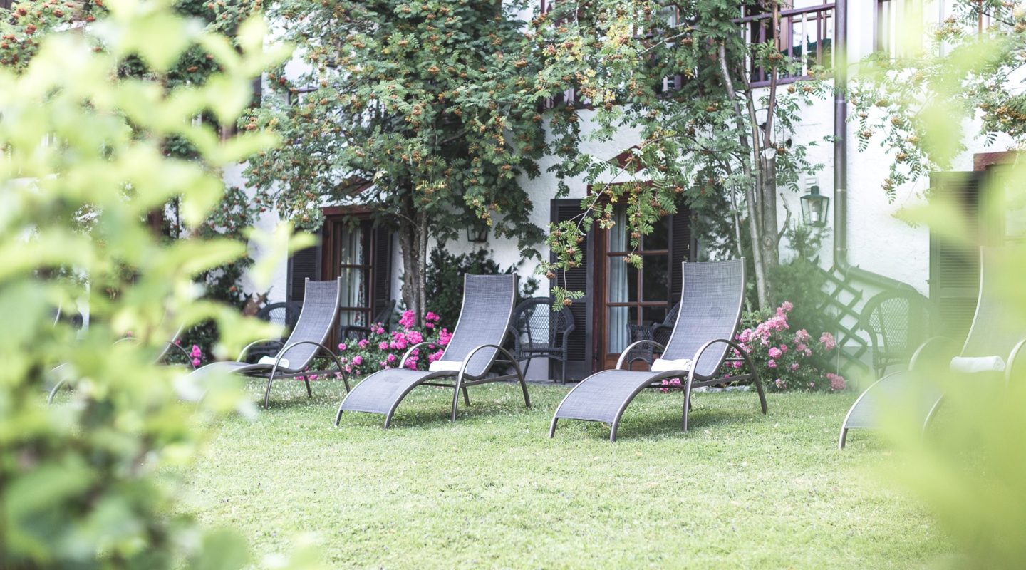 Three grey lounge chairs sit on a green lawn in front of a white building with trees and pink flowers.