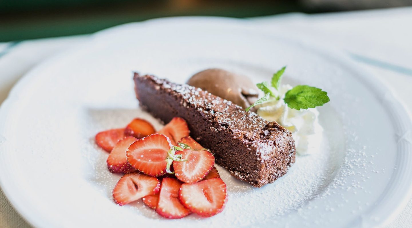 A slice of chocolate cake on a white plate with fresh sliced strawberries, whipped cream, and chocolate ice cream.