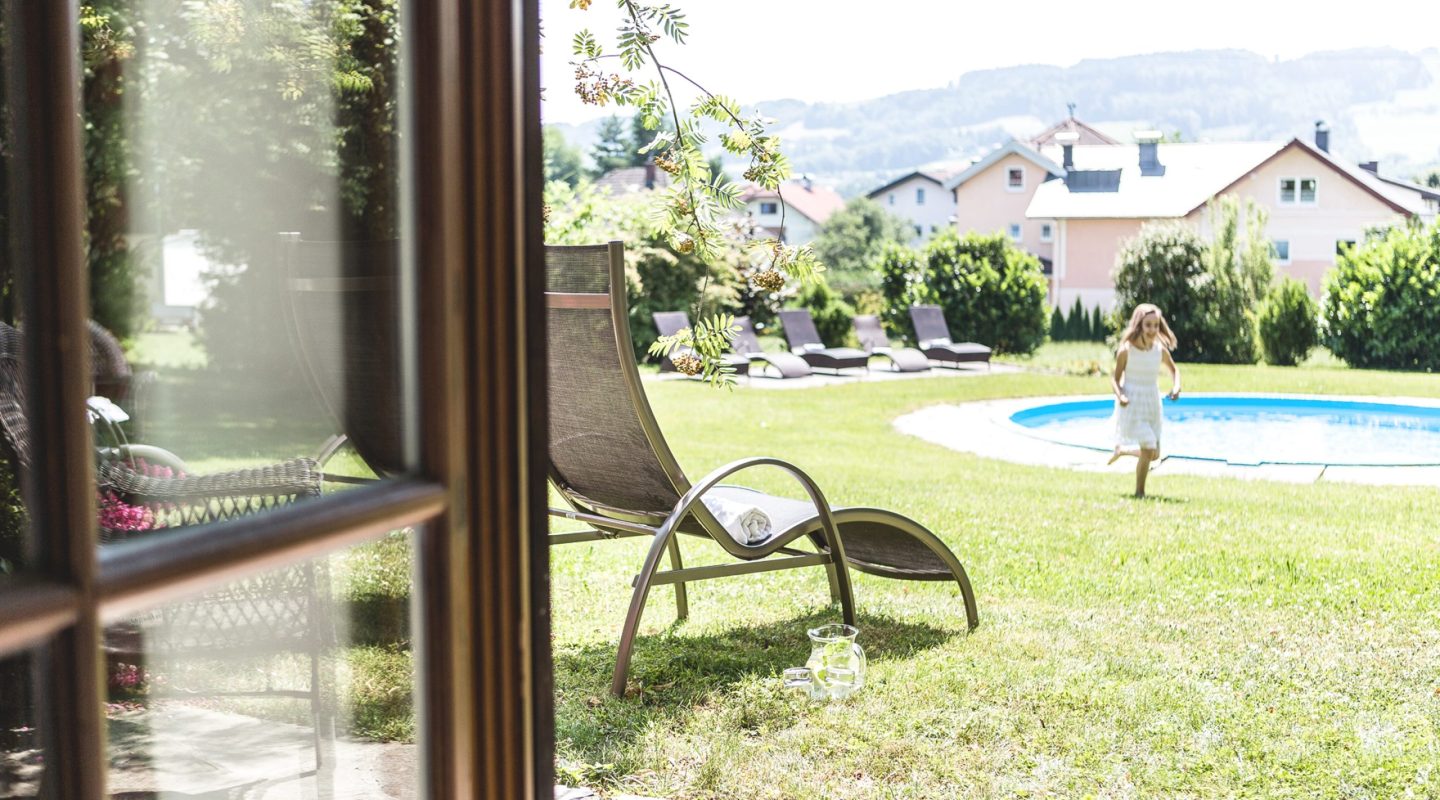 A young girl runs across a sunny green lawn near a swimming pool, seen from a doorway beside a lounge chair.