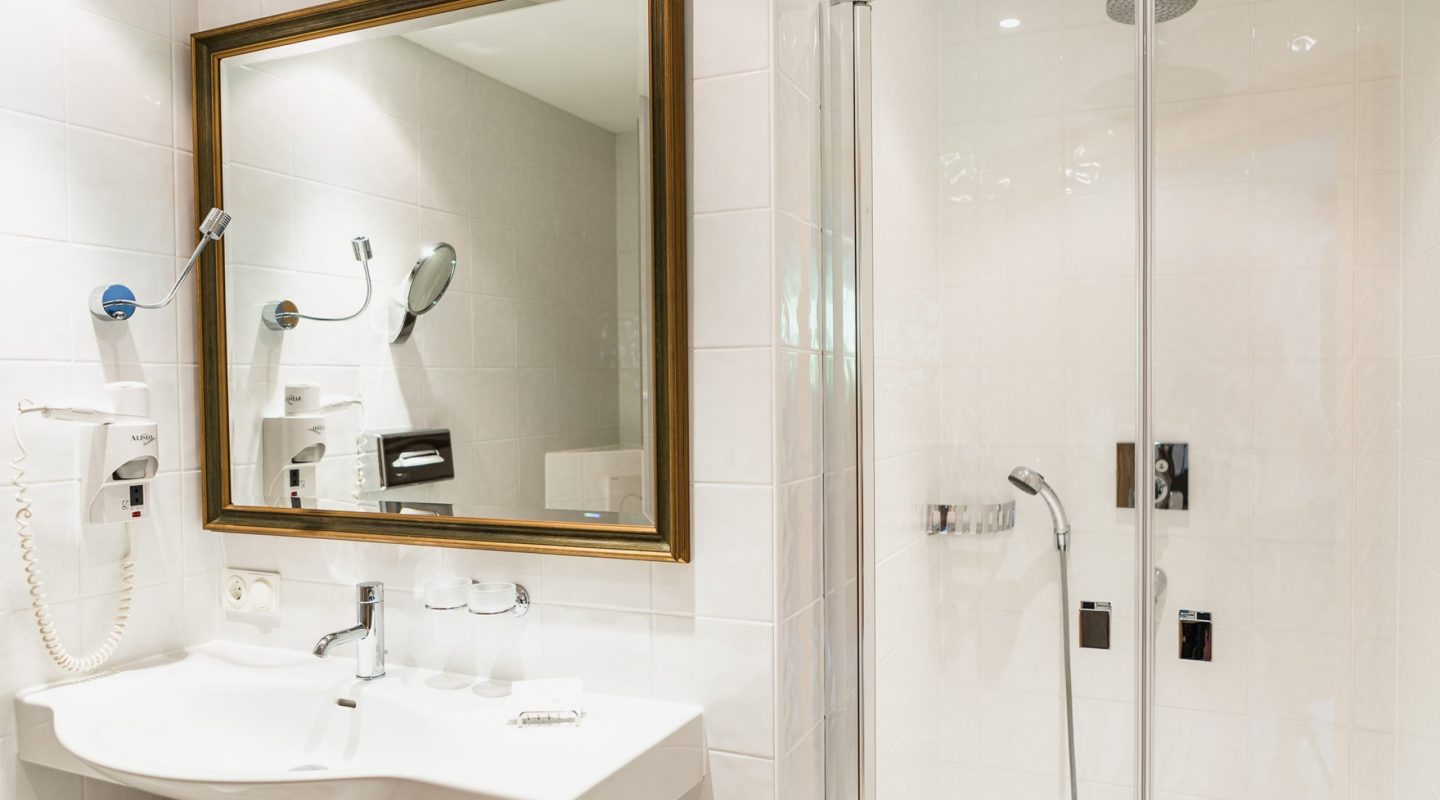 Modern hotel bathroom with a white ceramic sink, large gold-framed mirror, and a glass-enclosed walk-in shower.