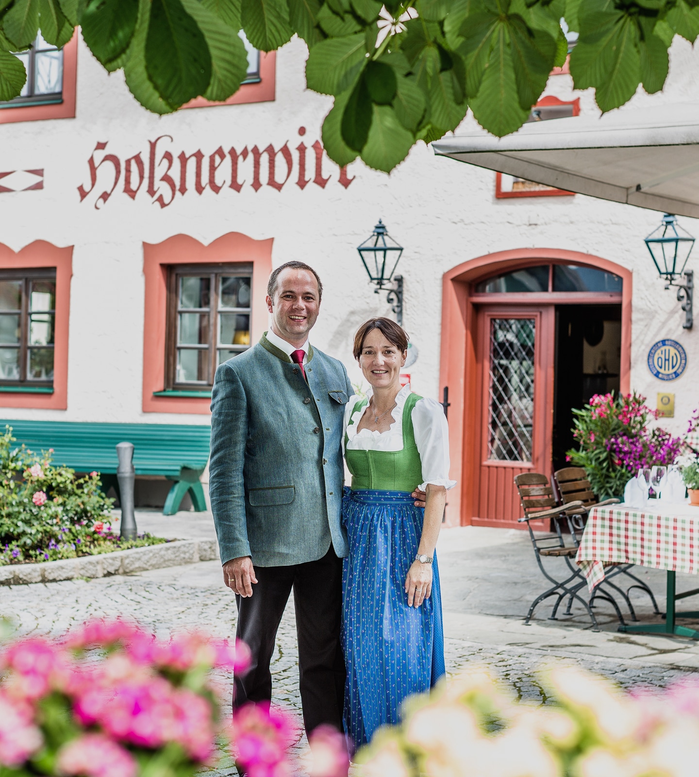 Ein lächelndes Paar in Tracht vor dem Gasthof Holznerwirt in Österreich.