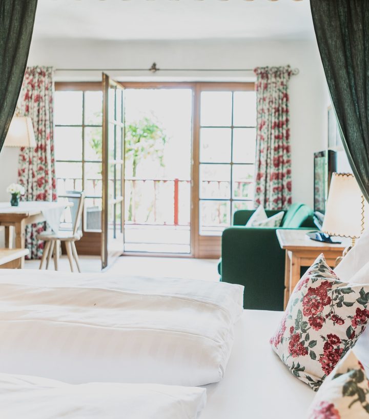 Gemütliches Hotelzimmer mit Bett im Vordergrund, Blick auf Sitzecke und offene Balkontür im Hintergrund.