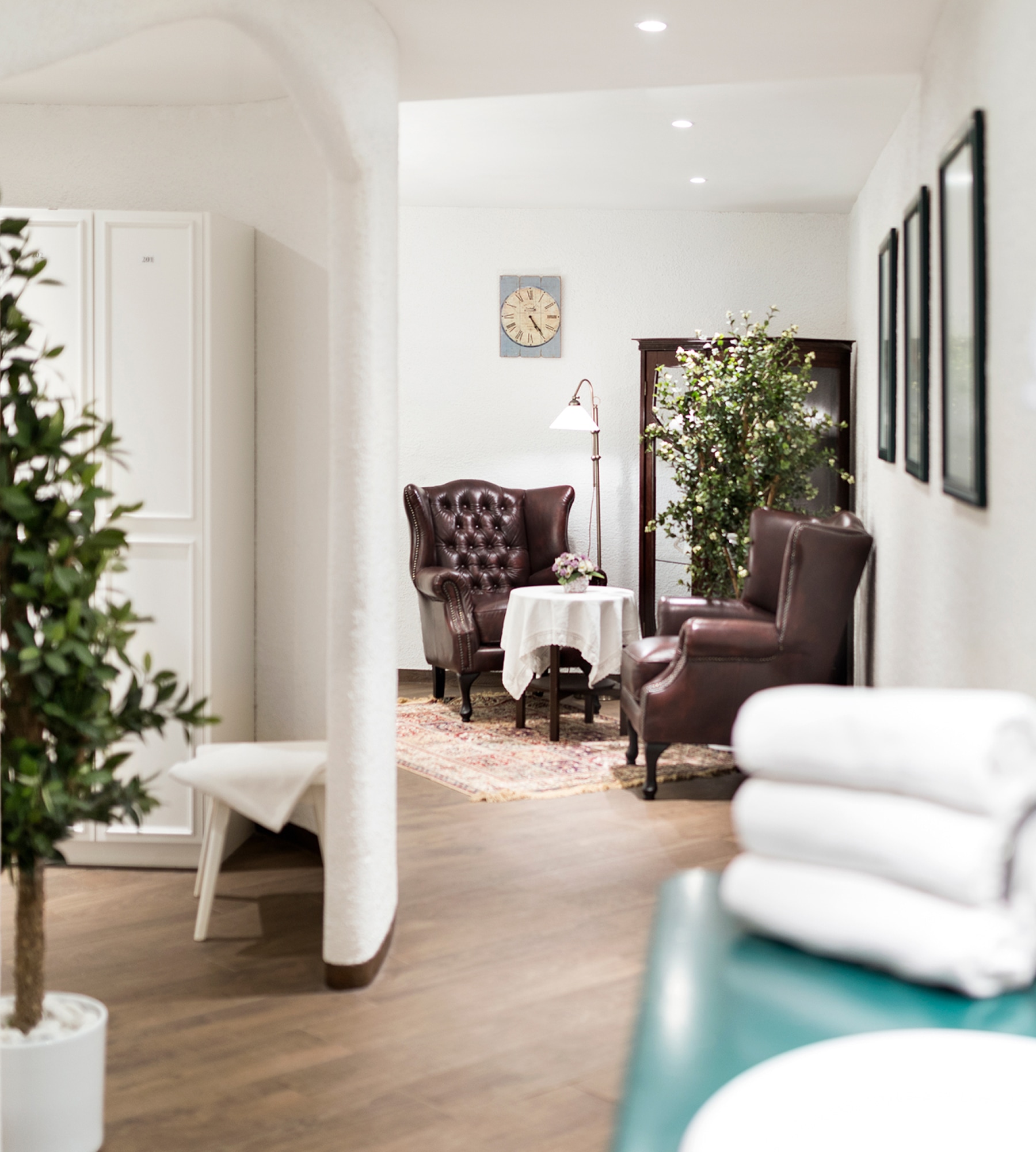 A cozy spa waiting area with tufted leather armchairs, a small table, a plant, and a stack of white towels.